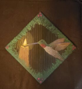 a small black canvas turned sideways to be diamond shaped, on it via neaon and metallic watercolours is a rubzthroated hummingbird with itäs beak touching the flame of a candle, behind is thin metal bars. around the border is red berries at each corner and on all around green foliage.
