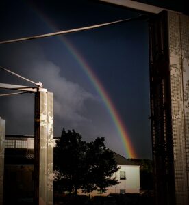 in the background a rainbow splits the sky into smooth dark on the right and a cloud on bright sky on the left. the midground has a white building with a black tree. the foreground has decorative white pillars with connecting poles.