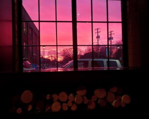 in the foreground are fireplace logs stacked along the bottom, above is a window with black grating, outside is a parking lot, a pale van parked near teh window, a building to the left side, telephone poles to the right, trees in teh distance, all lit by a vibrant pinkish red sunset 